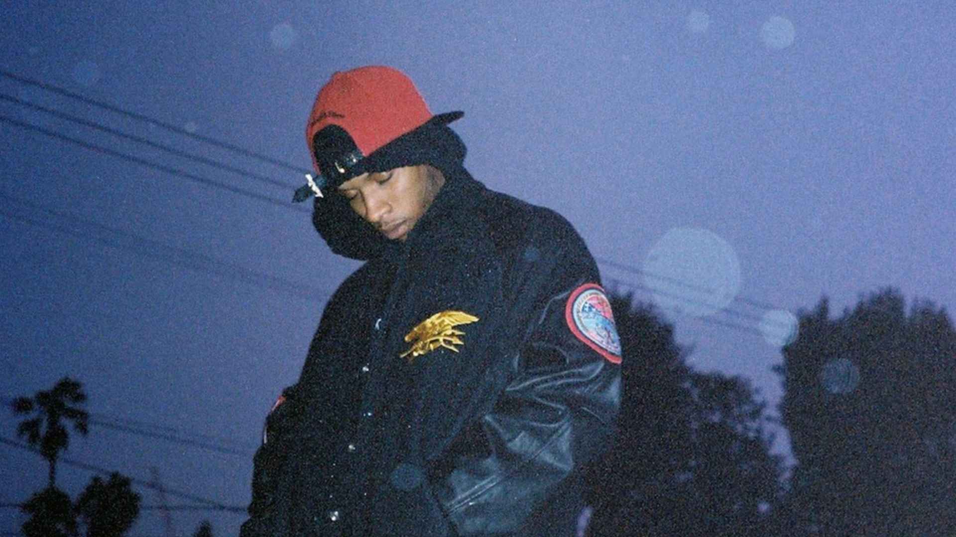 Tory Lanez poses outdoors at dusk in a black jacket and red cap, with palm trees in the background.