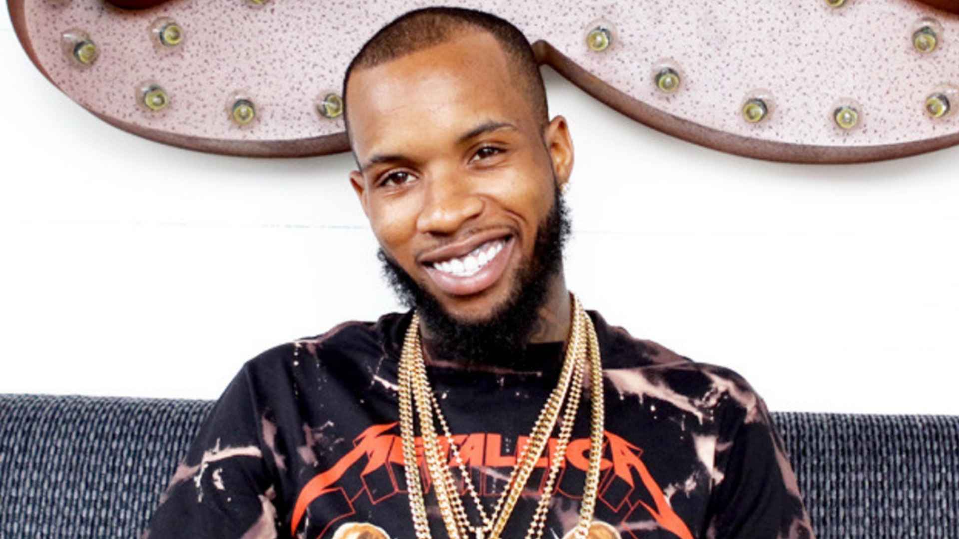 Tory Lanez smiles at the camera while seated indoors, wearing gold chains and a black graphic shirt.