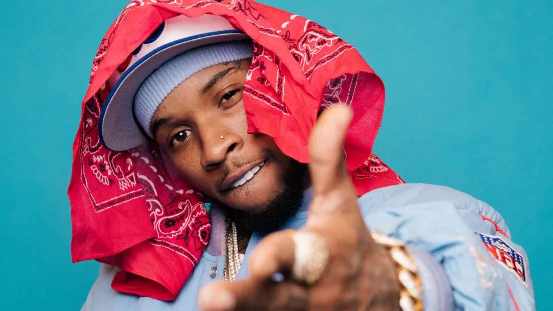 Tory Lanez poses against a blue backdrop, wearing a red bandana, cap, blue jacket, and gold jewelry.
