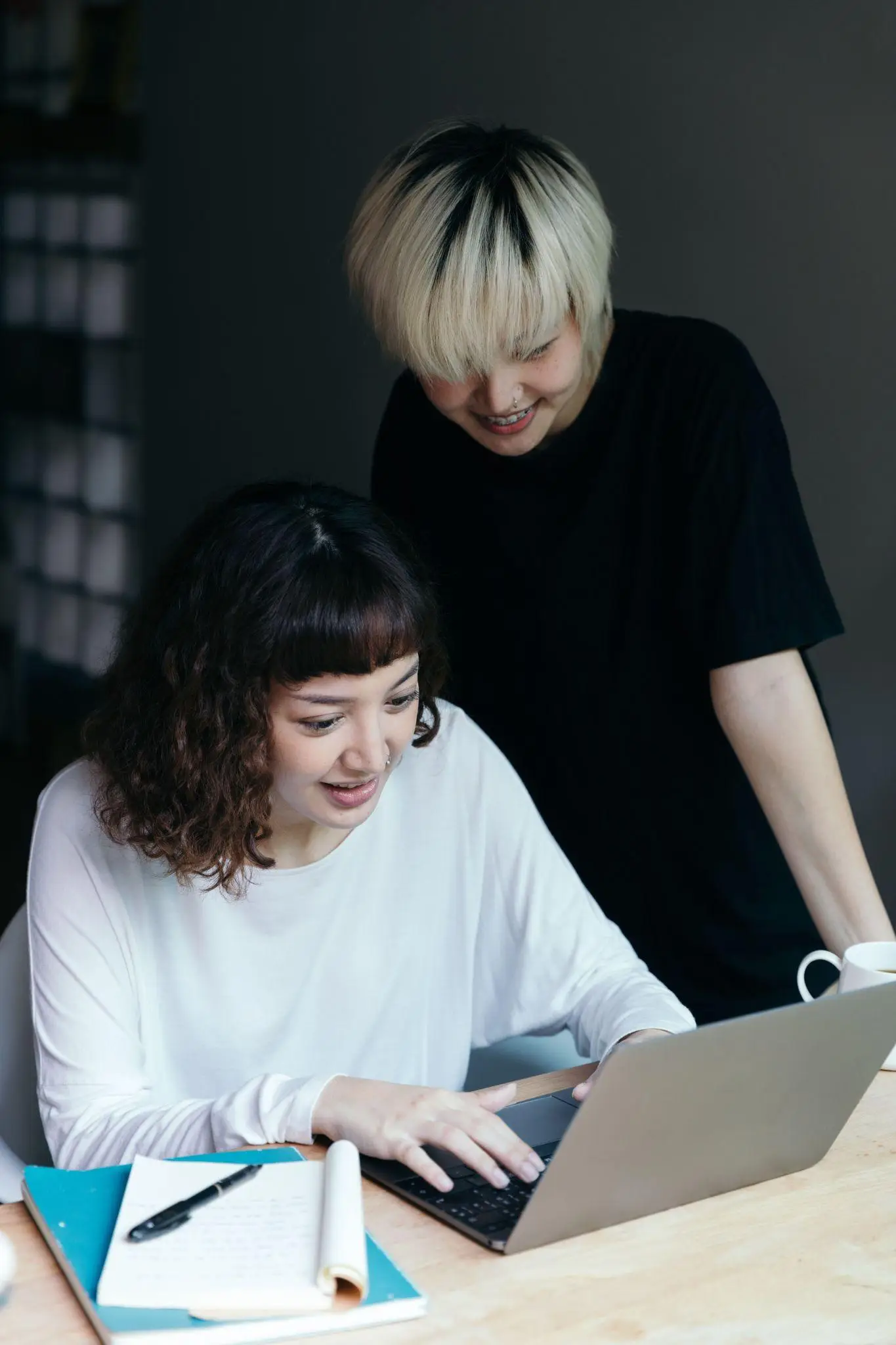 Two people using a laptop at a wooden table with notebooks and a coffee mug
