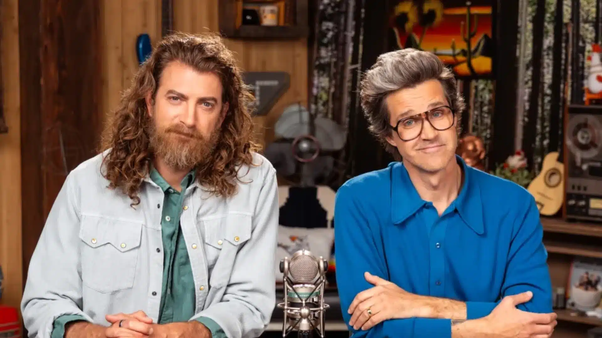 Two male YouTube hosts seated at a studio desk with a microphone between them