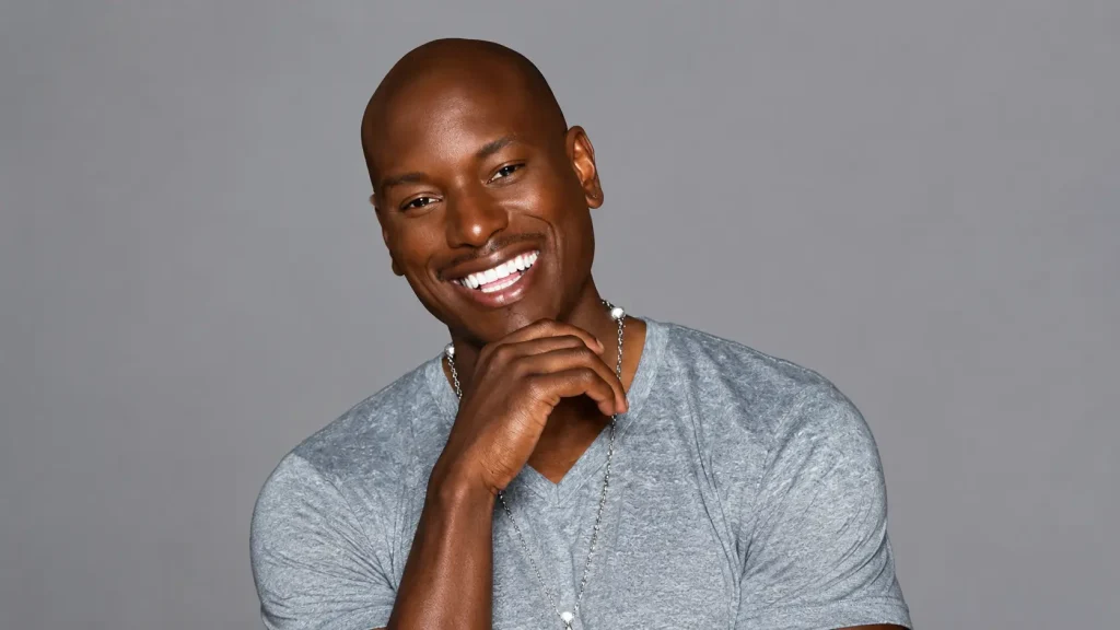 Tyrese Gibson wearing a gray V neck shirt and chain necklace posing with hand near chin against a studio background