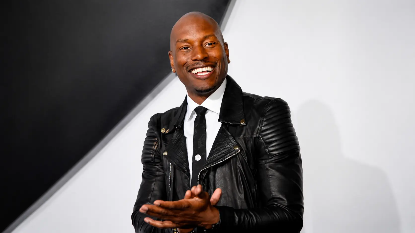 Tyrese Gibson wearing a black leather jacket and tie posing against a modern black and white backdrop at event