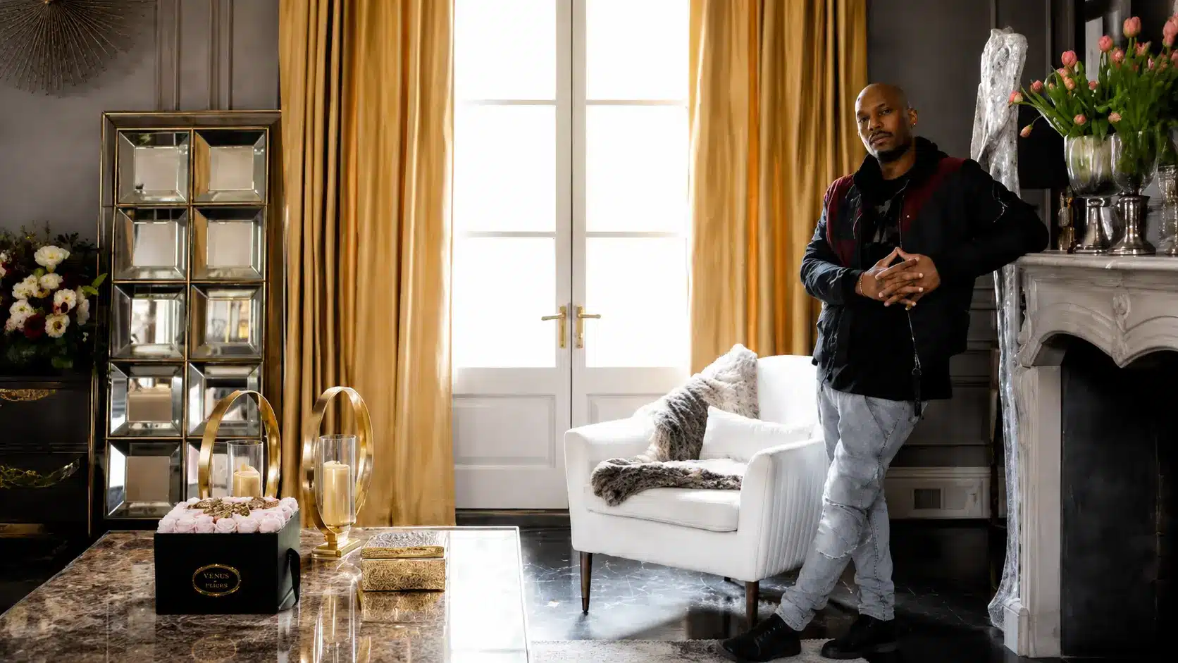 Luxury living room with tyrese gibson standing near fireplace featuring beige curtains white chair and refined interior decor