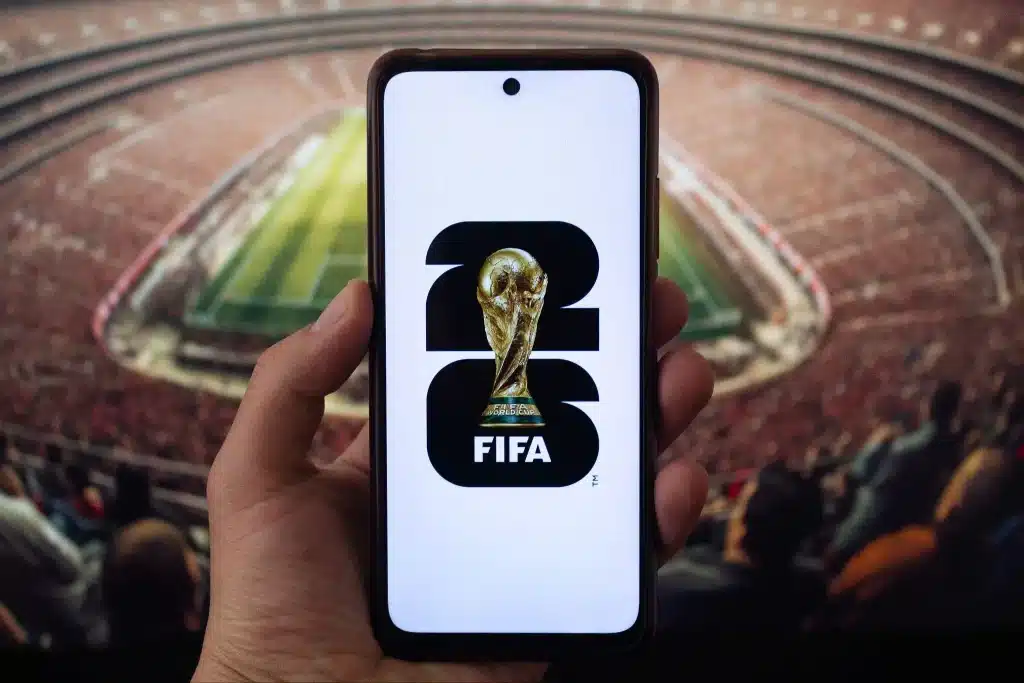 Hand holding smartphone displaying FIFA 2026 logo in front of large stadium crowd