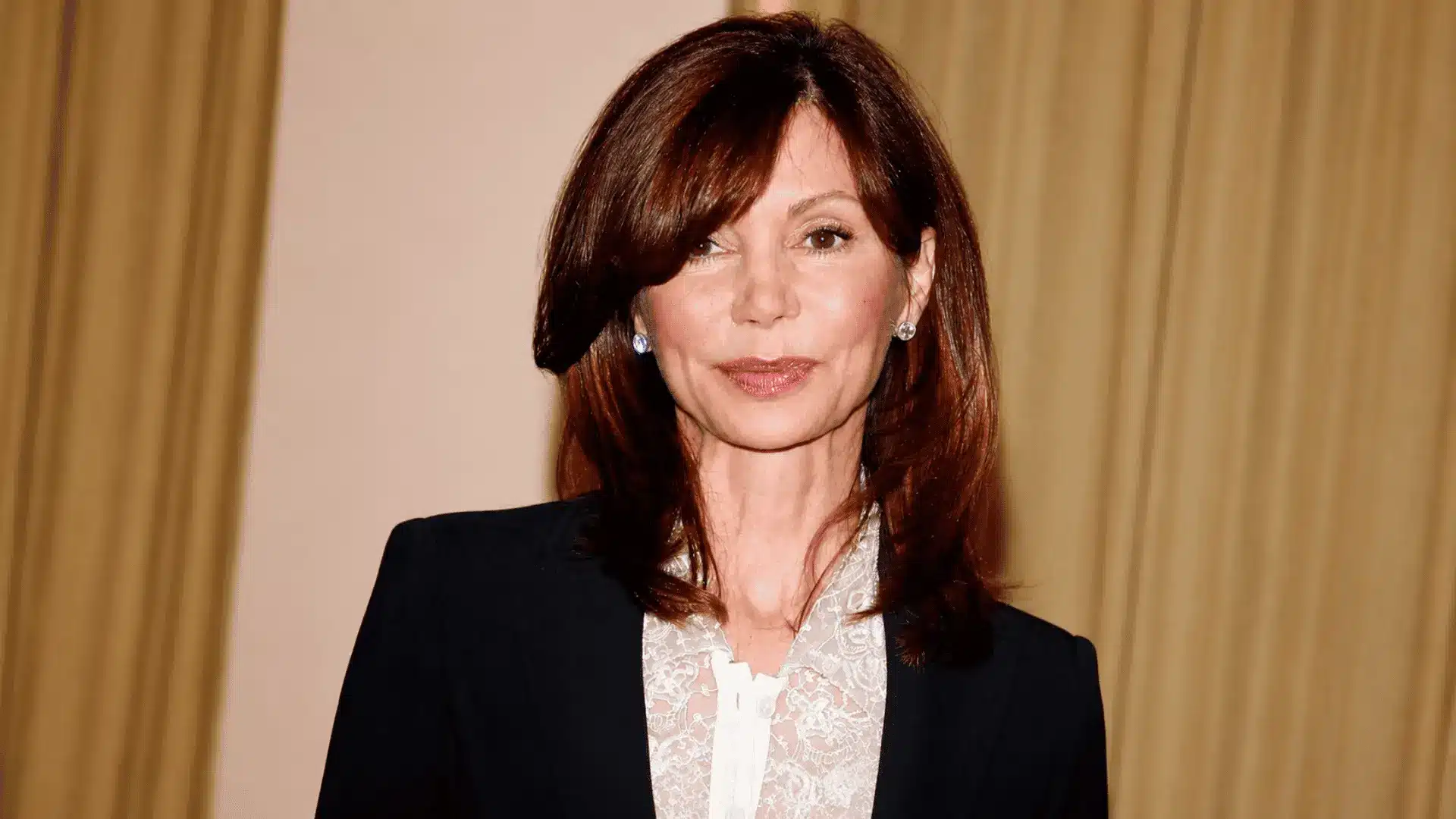 Victoria Principal wearing a black blazer and white lace blouse against a neutral indoor background