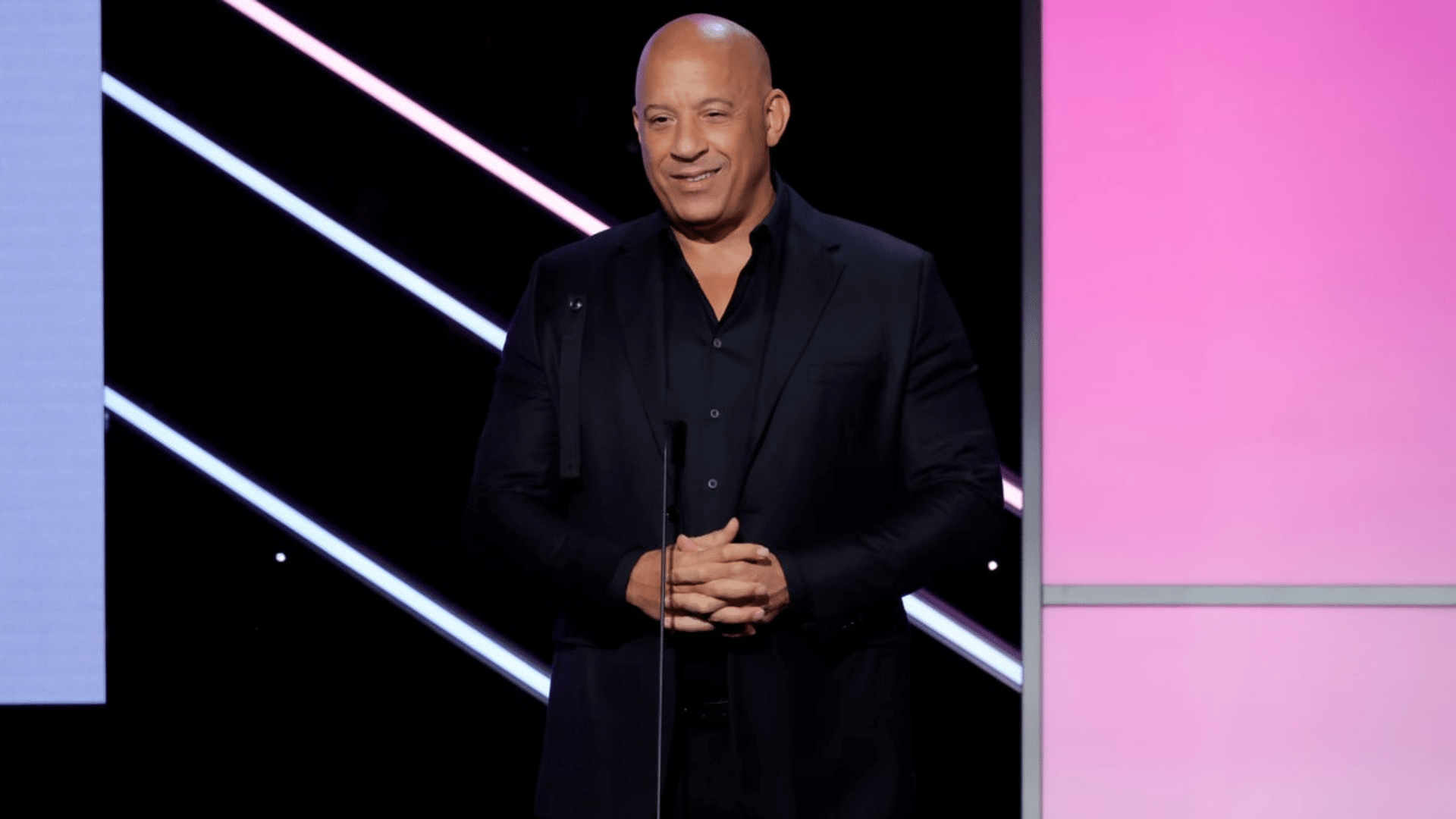 Vin Diesel wearing a black suit while speaking on stage during a public event with a modern illuminated background