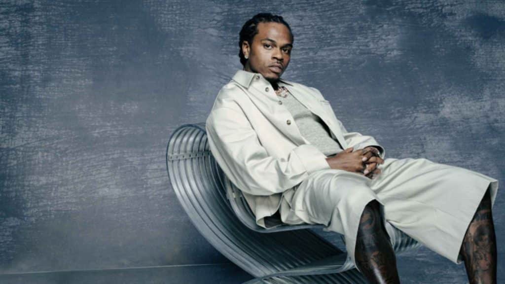 Gunna in a white outfit seated on a curved metal chair against a textured blue-gray backdrop.