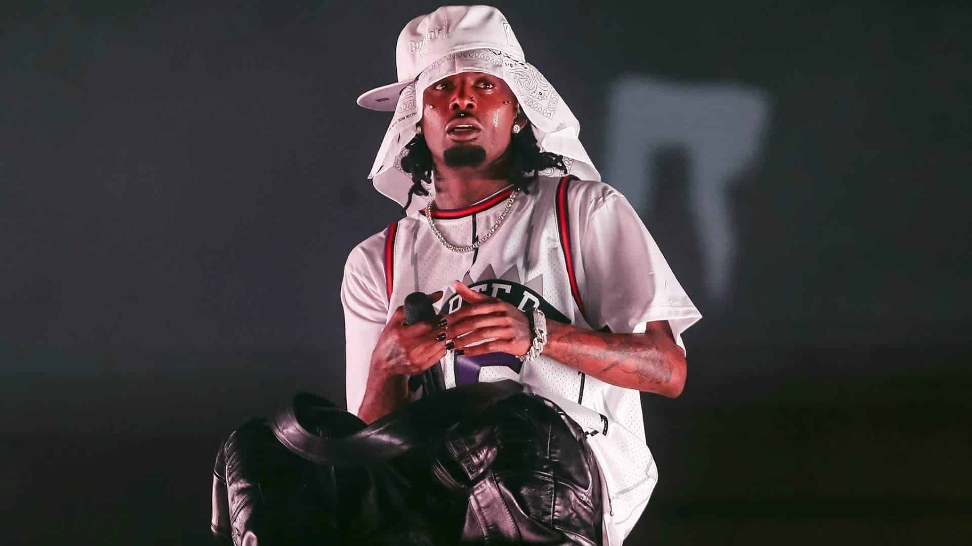 Musician Playboi Carti performing on stage in a white hat, bandana, jersey, and leather pants.