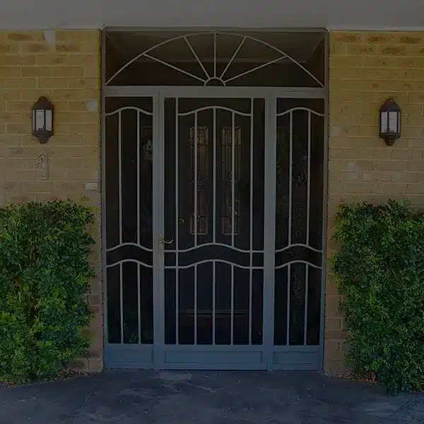 Screen door with decorative metalwork against brick wall flanked by lanterns and greenery