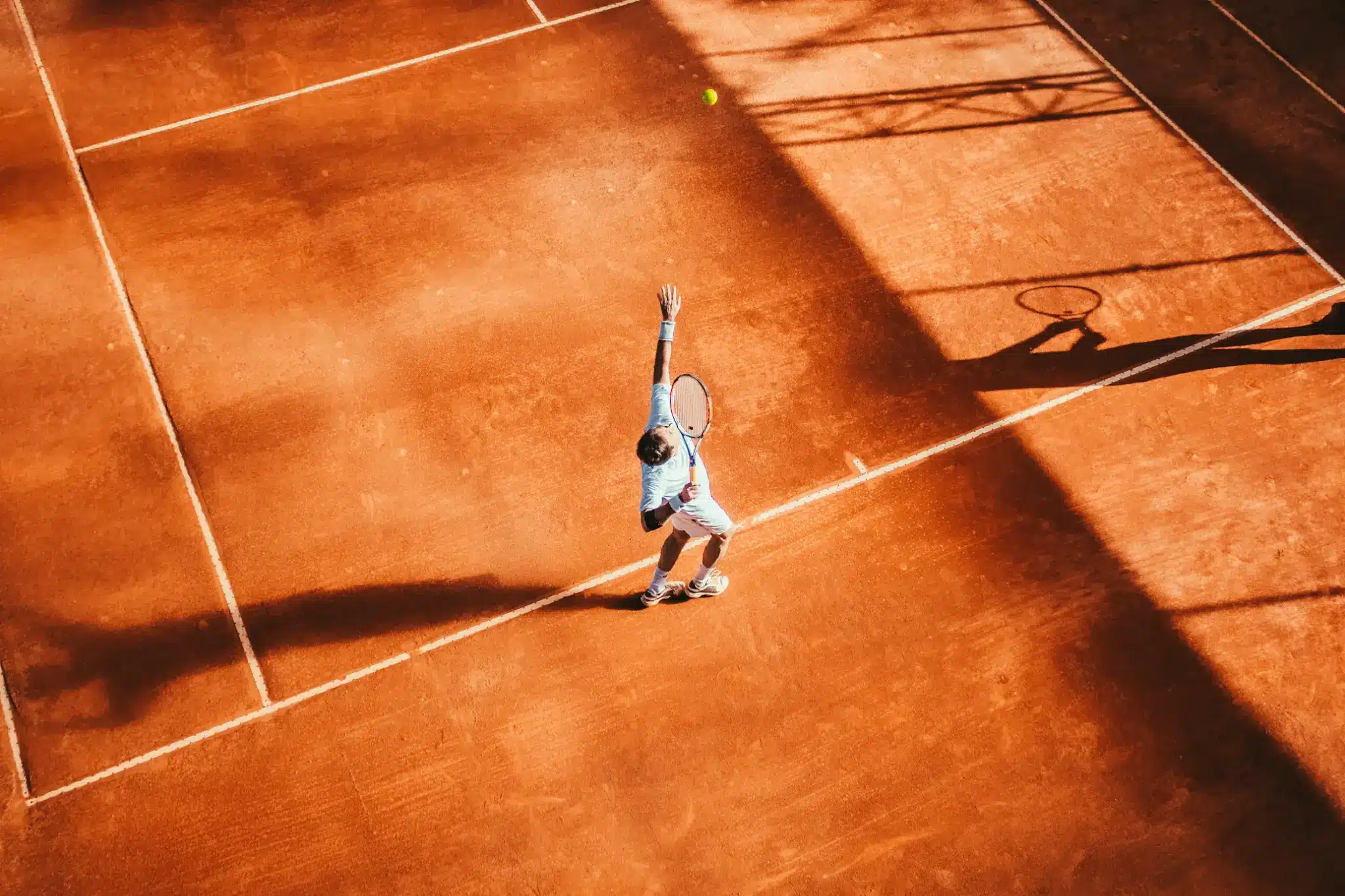 Tennis player preparing to serve on clay court under daylight shadows