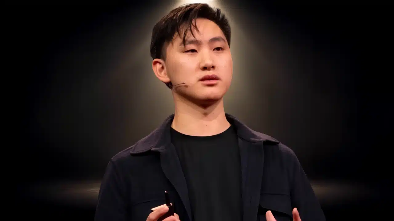 Young man in a black jacket speaking under a spotlight during a presentation.