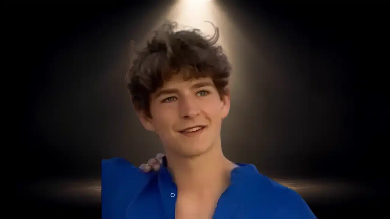 Young man with dark hair smiling in a blue shirt against a dark background.