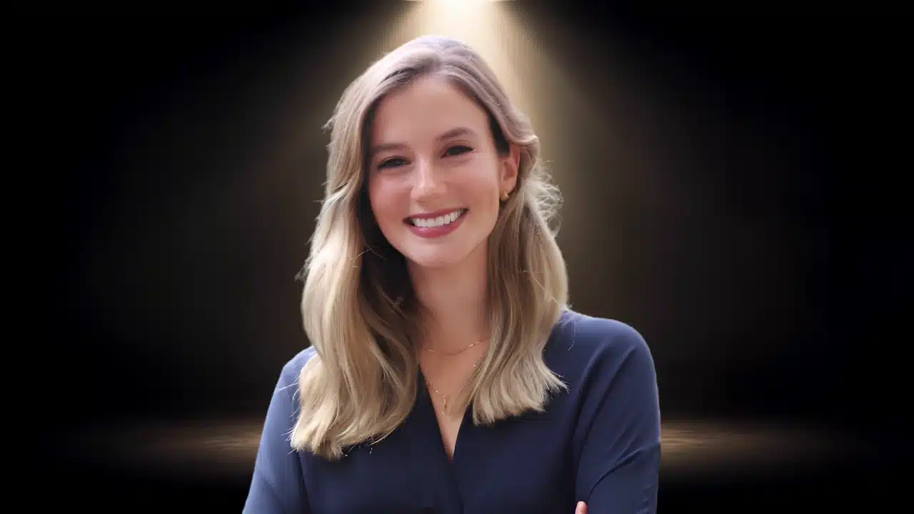Young woman with blonde hair smiling warmly in a navy blue top against a dark background.