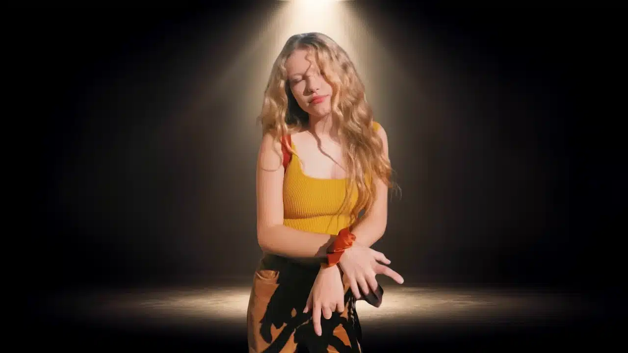 Young woman with long blonde hair posing under a spotlight in a yellow top.