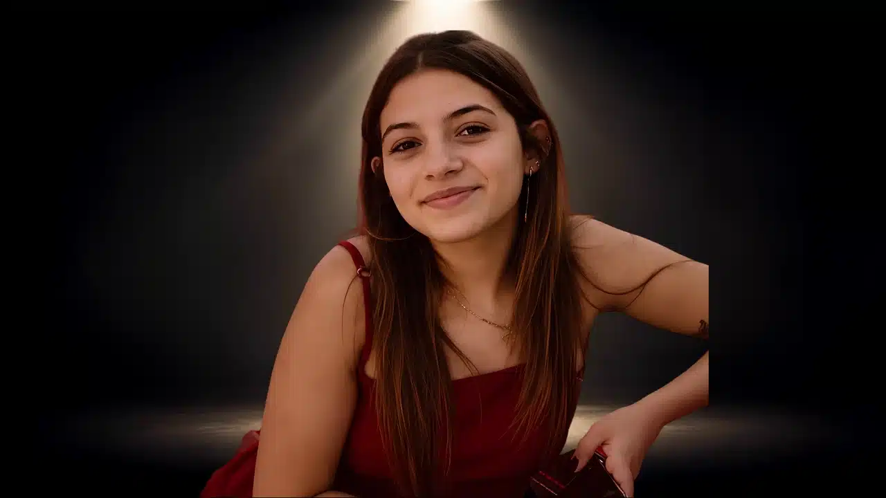 Young woman with long brown hair smiling in a red top under soft overhead lighting.