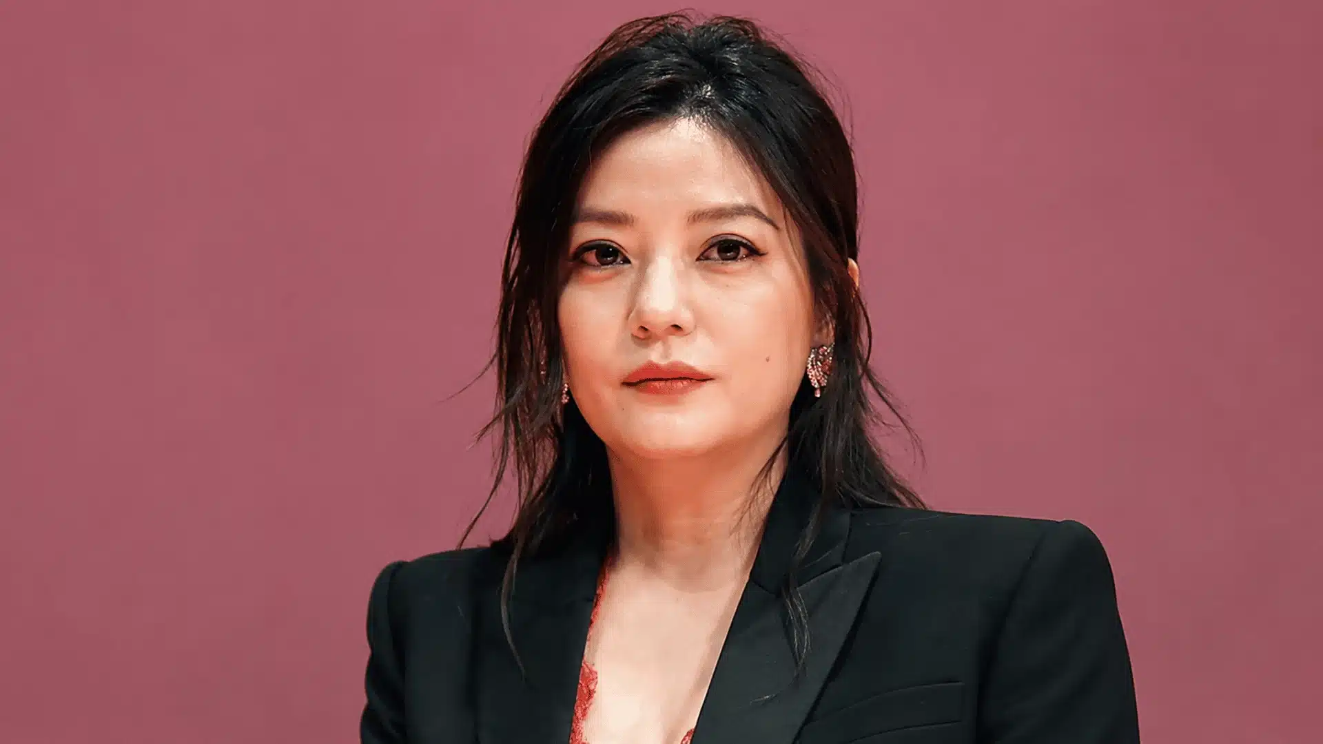 Zhao Wei wearing a black blazer and earrings while posing against a muted pink background