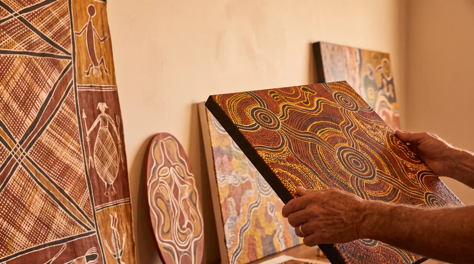 Hands examining Aboriginal art painting in warm-toned studio with multiple artworks displayed