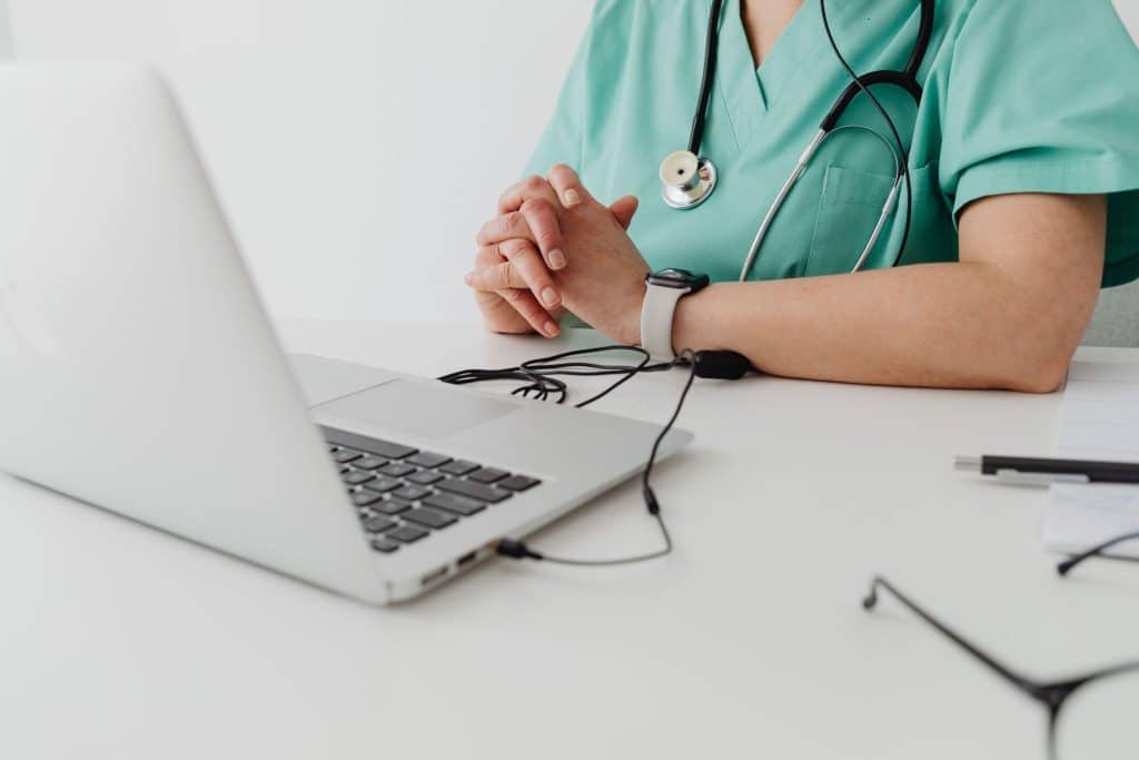 Medical professional in scrubs with stethoscope sitting at desk with open laptop