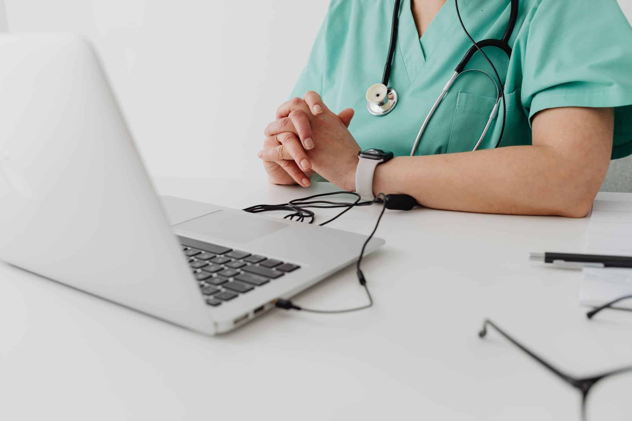 Medical professional in scrubs with stethoscope sitting at desk with open laptop