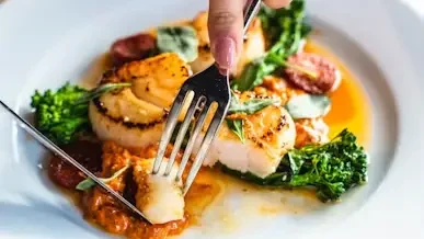 Person cutting into seared scallops with broccoli and tomatoes on white plate in natural light
