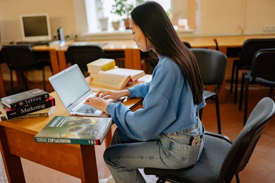 Person typing on laptop at wooden desk in bright library setting with books nearby