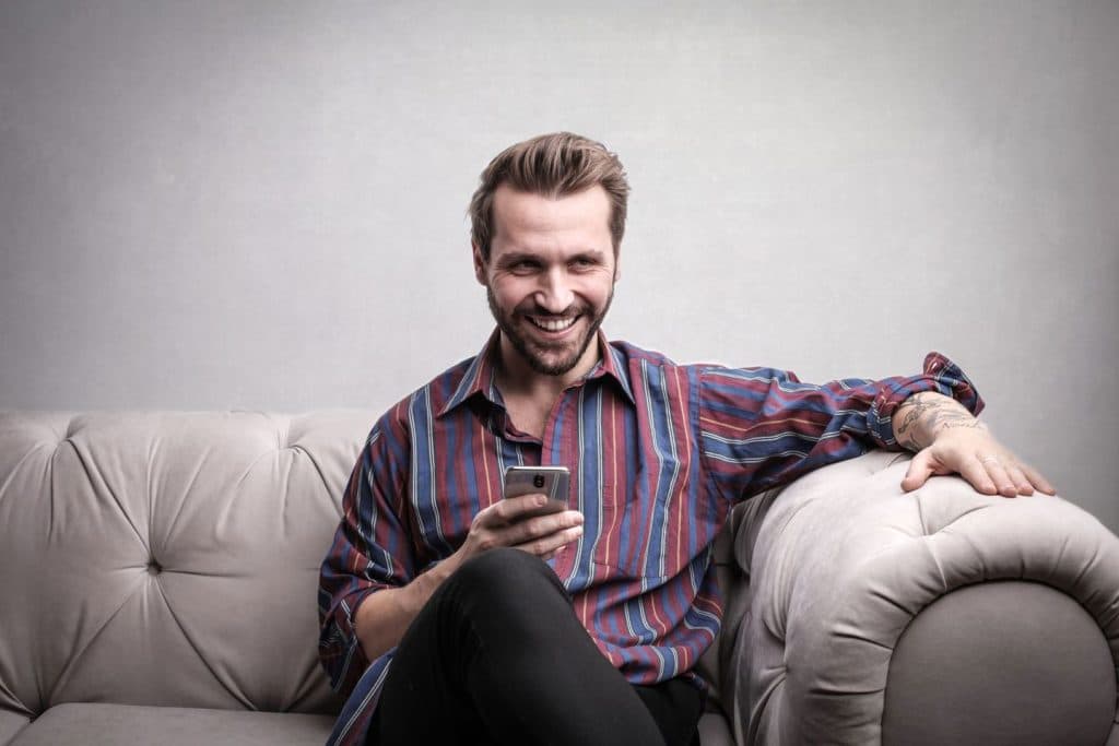 Man smiling while holding smartphone on light gray sofa indoors