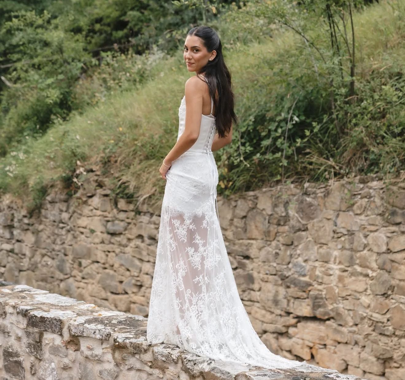 Woman in lace wedding dress standing on stone wall in lush outdoor setting
